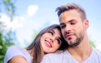 A young woman with long brown hair rests her head on a man's shoulder. Both are smiling slightly, wearing white shirts, and sitting outdoors against a blurred background of blue sky and greenery.