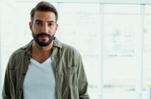 A man with a beard and short hair is standing in front of large windows. He is wearing a white T-shirt and an open olive-green button-up shirt. The background is bright and slightly blurred.