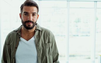 A man with a beard and short hair is standing in front of large windows. He is wearing a white T-shirt and an open olive-green button-up shirt. The background is bright and slightly blurred.