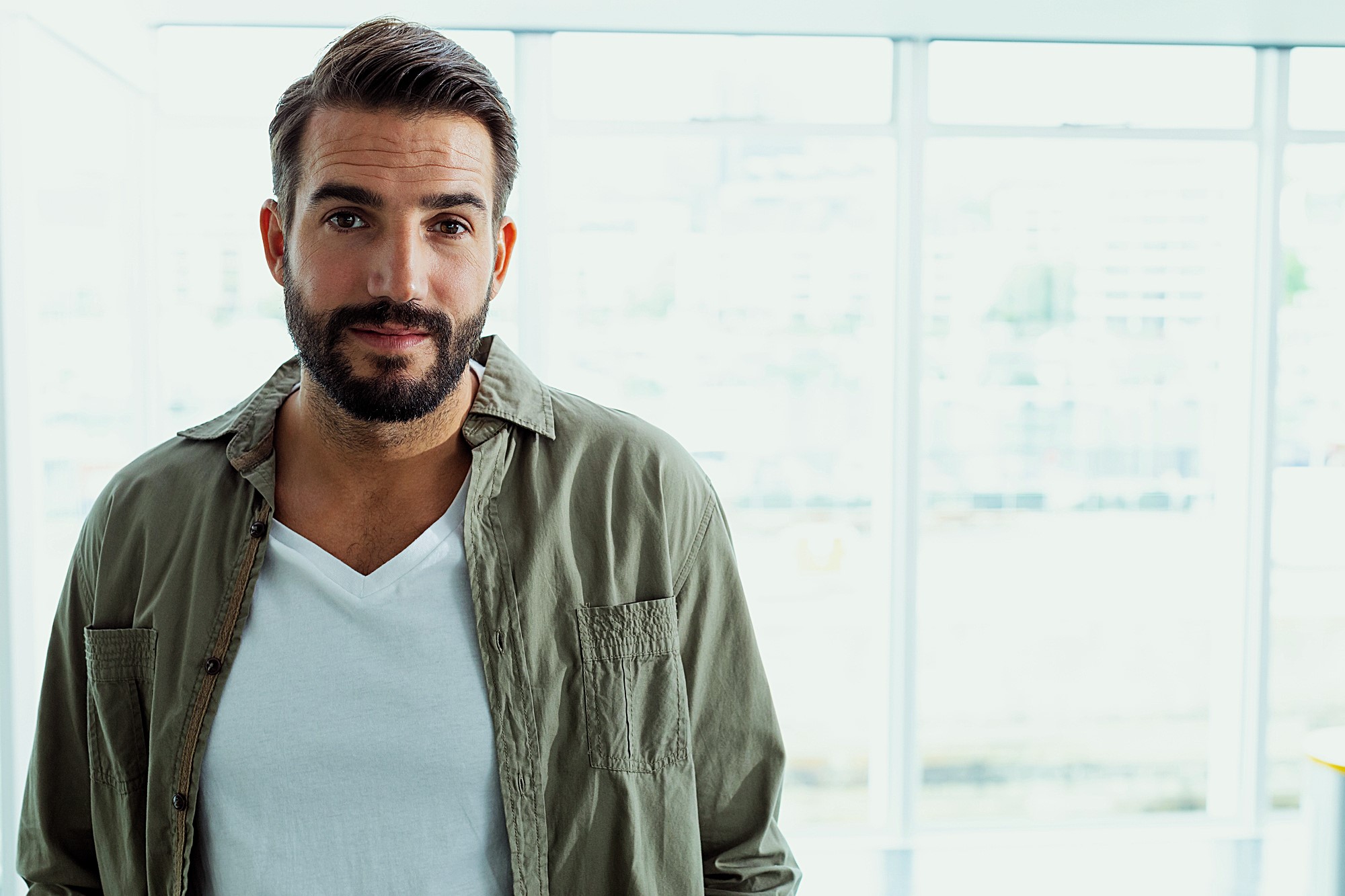 A man with a beard and short hair is standing in front of large windows. He is wearing a white T-shirt and an open olive-green button-up shirt. The background is bright and slightly blurred.