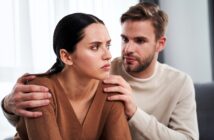 A man with a beard is gently holding a woman's shoulders. The woman, with long dark hair, looks away with a serious expression. They are sitting indoors, and the man appears to be speaking to her. Both are wearing casual sweaters.