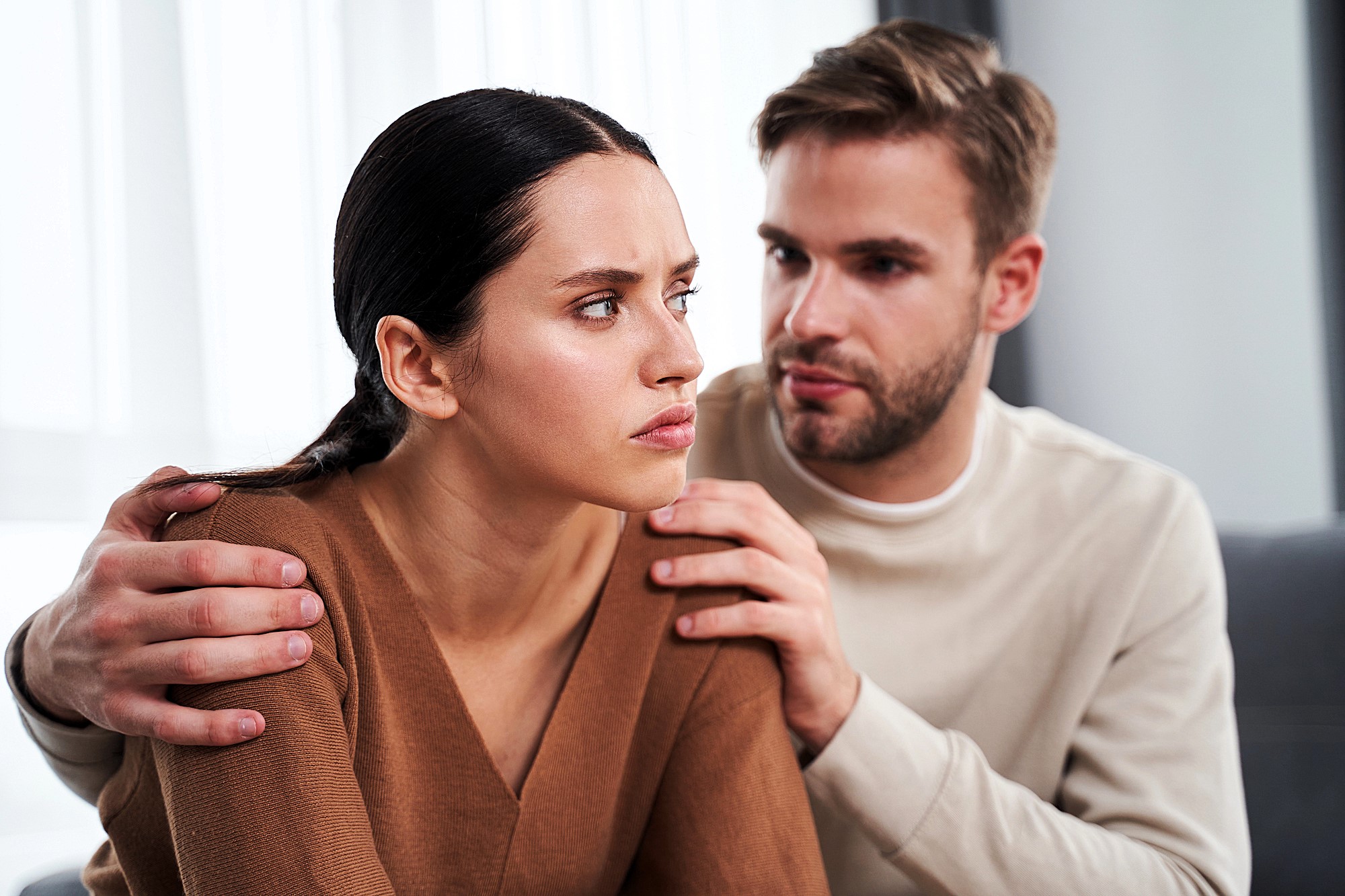 A man with a beard is gently holding a woman's shoulders. The woman, with long dark hair, looks away with a serious expression. They are sitting indoors, and the man appears to be speaking to her. Both are wearing casual sweaters.