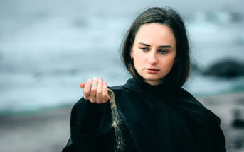 A woman with long dark hair is on a beach, wearing a black cloak. She looks thoughtfully at sand falling from her hand, with an overcast sky and ocean waves in the background.