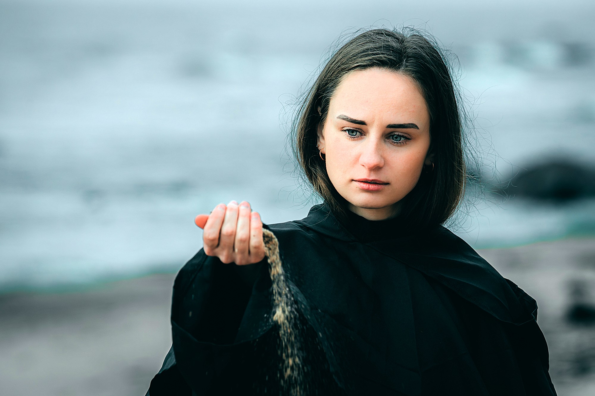 A woman with long dark hair is on a beach, wearing a black cloak. She looks thoughtfully at sand falling from her hand, with an overcast sky and ocean waves in the background.