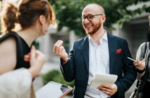 A bearded man in a suit and glasses holds a notepad and pen while smiling and talking to a woman. She has her back to the camera and holds a clipboard. Another person beside them holds a phone. Greenery is blurred in the background.