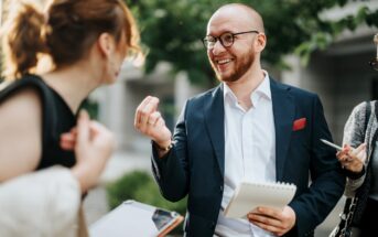 A bearded man in a suit and glasses holds a notepad and pen while smiling and talking to a woman. She has her back to the camera and holds a clipboard. Another person beside them holds a phone. Greenery is blurred in the background.