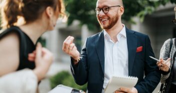 A bearded man in a suit and glasses holds a notepad and pen while smiling and talking to a woman. She has her back to the camera and holds a clipboard. Another person beside them holds a phone. Greenery is blurred in the background.