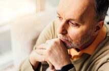 A man with a thoughtful expression looks into the distance, resting his chin on his hands. He has short hair, a beard, and is wearing a light-colored sweater over an orange shirt. Soft lighting creates a warm atmosphere.