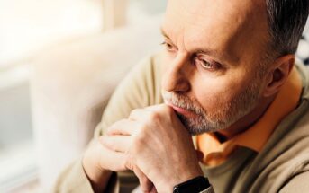 A man with a thoughtful expression looks into the distance, resting his chin on his hands. He has short hair, a beard, and is wearing a light-colored sweater over an orange shirt. Soft lighting creates a warm atmosphere.