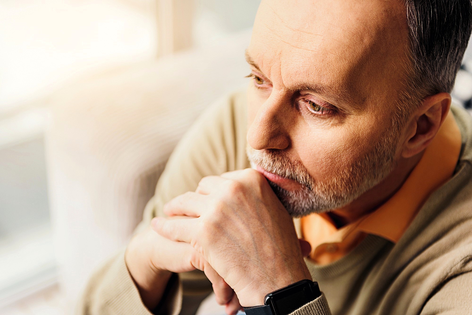 A man with a thoughtful expression looks into the distance, resting his chin on his hands. He has short hair, a beard, and is wearing a light-colored sweater over an orange shirt. Soft lighting creates a warm atmosphere.