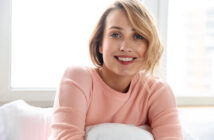 A smiling woman with short blonde hair leans against a pillow, wearing a light pink sweater. She is sitting in a brightly lit room with large windows in the background.