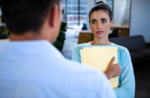 A woman holding a stack of yellow folders looks concerned while talking to a man whose back faces the viewer. They are in a modern office setting with a couch and large windows in the background.