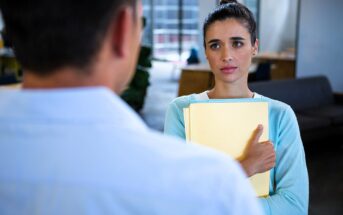 A woman holding a stack of yellow folders looks concerned while talking to a man whose back faces the viewer. They are in a modern office setting with a couch and large windows in the background.