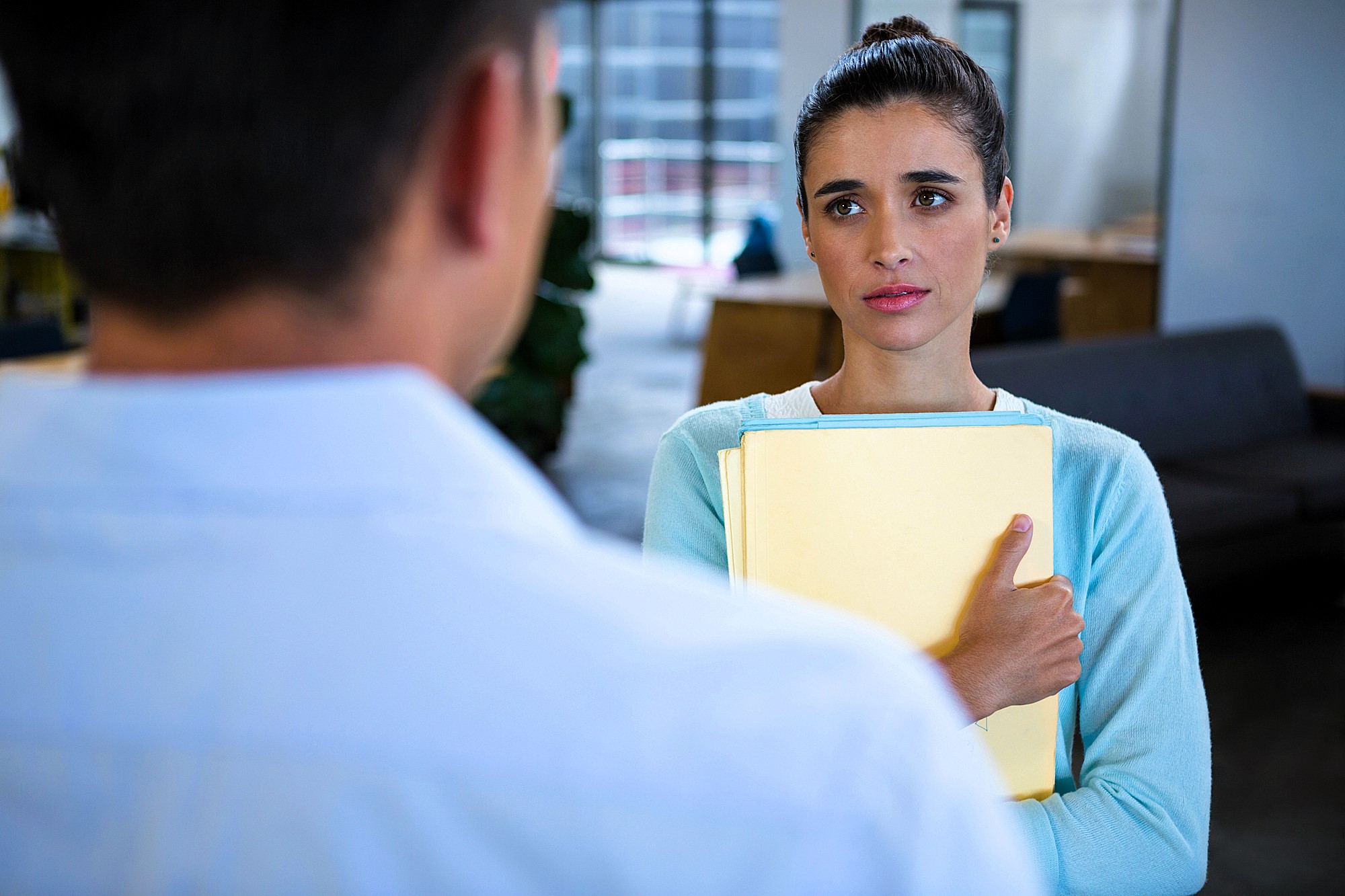 A woman holding a stack of yellow folders looks concerned while talking to a man whose back faces the viewer. They are in a modern office setting with a couch and large windows in the background.