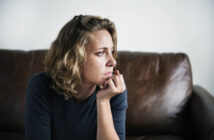 A woman with curly blonde hair wearing a dark sweater sits thoughtfully on a brown leather couch, resting her chin on her hand. She gazes to the side with a contemplative expression. The background is plain and neutral.