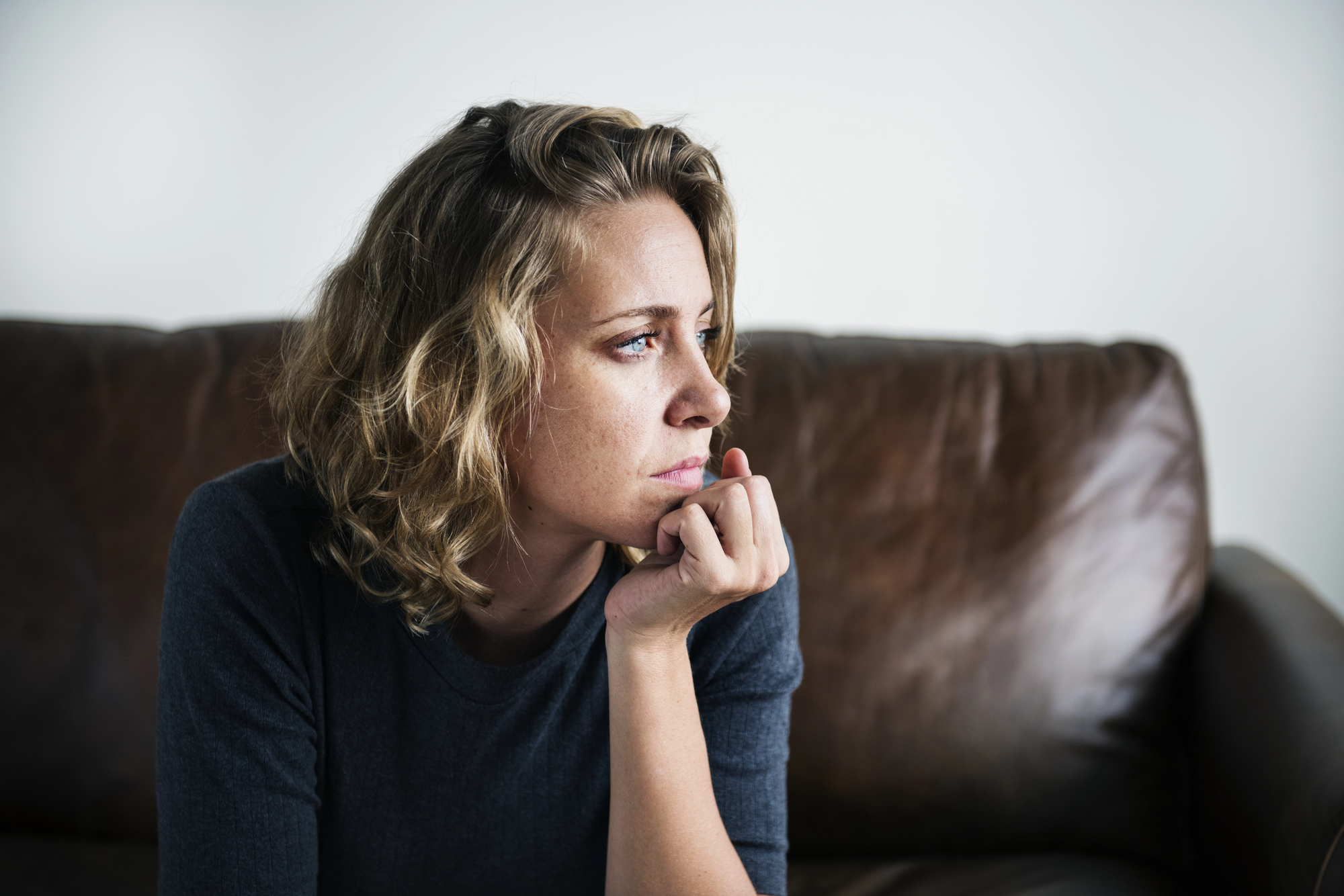 A woman with curly blonde hair wearing a dark sweater sits thoughtfully on a brown leather couch, resting her chin on her hand. She gazes to the side with a contemplative expression. The background is plain and neutral.