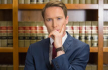 A person in a suit with a thoughtful expression stands in front of a wooden bookshelf filled with legal books.