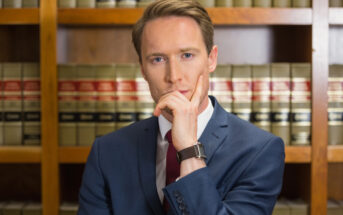 A person in a suit with a thoughtful expression stands in front of a wooden bookshelf filled with legal books.