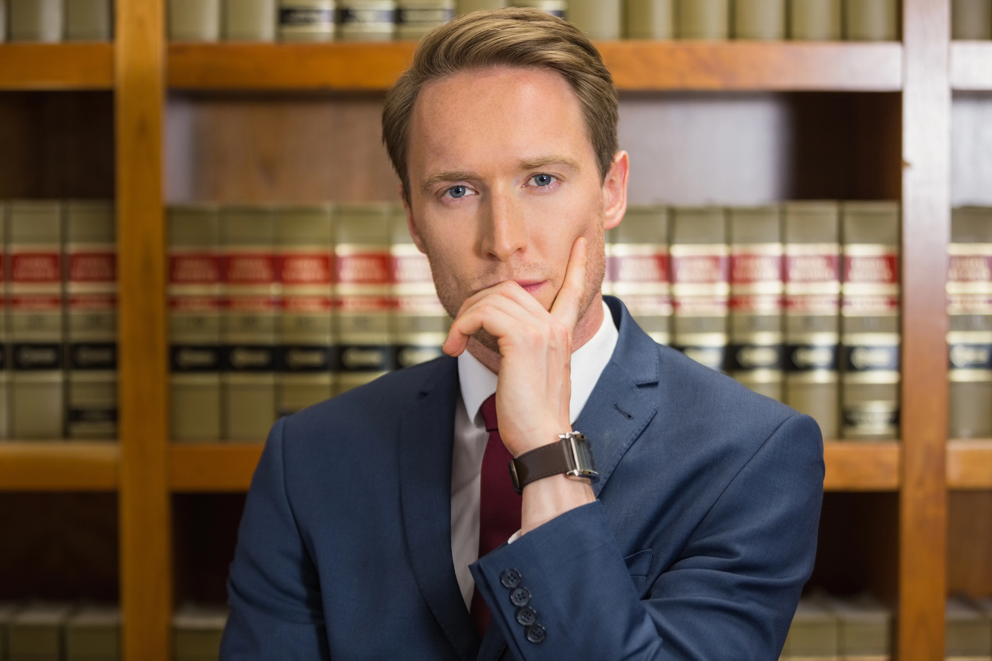 A person in a suit with a thoughtful expression stands in front of a wooden bookshelf filled with legal books.