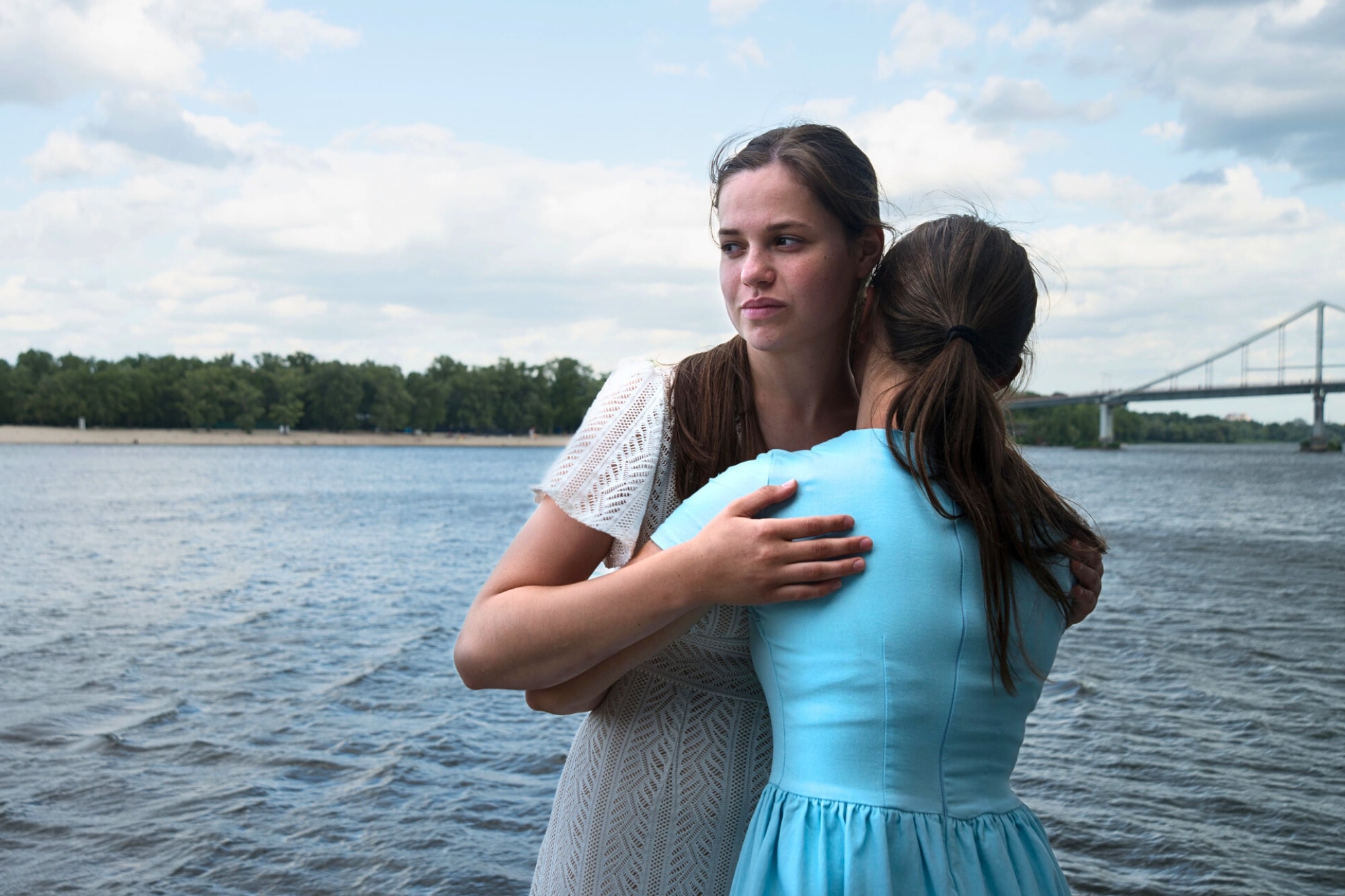 Two women embracing by a river under a partly cloudy sky. One woman faces the camera with a thoughtful expression, wearing a white dress, while the other, in a blue dress, has her back to the camera. A bridge and trees are visible in the background.