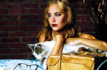 A woman with long hair sits at a table in front of a brick wall, holding a white and gray cat. She gazes thoughtfully into the distance. On the table are a pair of glasses and a tan leather bag.