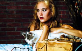 A woman with long hair sits at a table in front of a brick wall, holding a white and gray cat. She gazes thoughtfully into the distance. On the table are a pair of glasses and a tan leather bag.