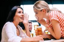 Two women are sitting at a table with glasses of beer, engaged in a lively conversation. One is whispering to the other, while the other looks surprised. The scene is set in a bright, casual setting with people in the background.