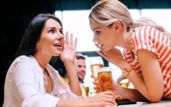 Two women are sitting at a table with glasses of beer, engaged in a lively conversation. One is whispering to the other, while the other looks surprised. The scene is set in a bright, casual setting with people in the background.