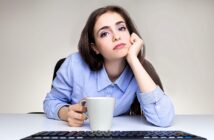 A woman with long brown hair rests her chin on her hand, looking at the camera with a neutral expression. She is holding a white mug and sits at a desk with a keyboard visible in the foreground. She wears a light blue button-up shirt.