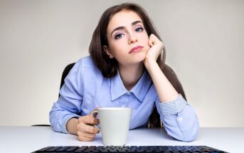 A woman with long brown hair rests her chin on her hand, looking at the camera with a neutral expression. She is holding a white mug and sits at a desk with a keyboard visible in the foreground. She wears a light blue button-up shirt.