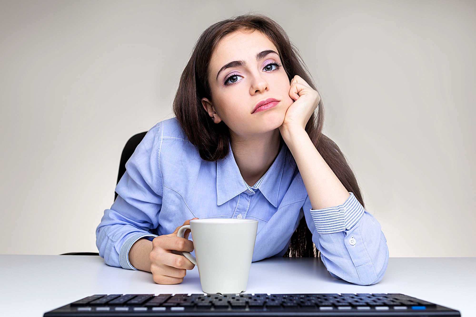 A woman with long brown hair rests her chin on her hand, looking at the camera with a neutral expression. She is holding a white mug and sits at a desk with a keyboard visible in the foreground. She wears a light blue button-up shirt.