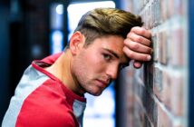 A person with short brown hair leans against a brick wall, wearing a red and gray shirt. They have a contemplative expression and are looking directly at the camera. The background is softly blurred.