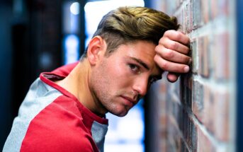 A person with short brown hair leans against a brick wall, wearing a red and gray shirt. They have a contemplative expression and are looking directly at the camera. The background is softly blurred.