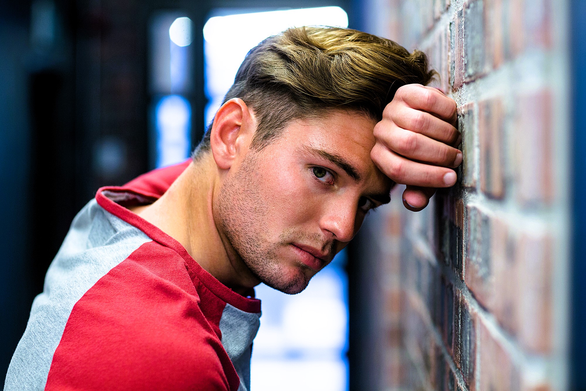 A person with short brown hair leans against a brick wall, wearing a red and gray shirt. They have a contemplative expression and are looking directly at the camera. The background is softly blurred.