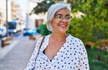 Smiling older woman with gray hair and glasses stands outdoors, wearing a white blouse with black patterns. The background features a tree and a blurred street scene.