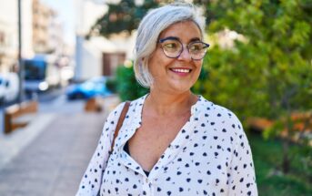 Smiling older woman with gray hair and glasses stands outdoors, wearing a white blouse with black patterns. The background features a tree and a blurred street scene.