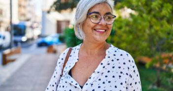 Smiling older woman with gray hair and glasses stands outdoors, wearing a white blouse with black patterns. The background features a tree and a blurred street scene.