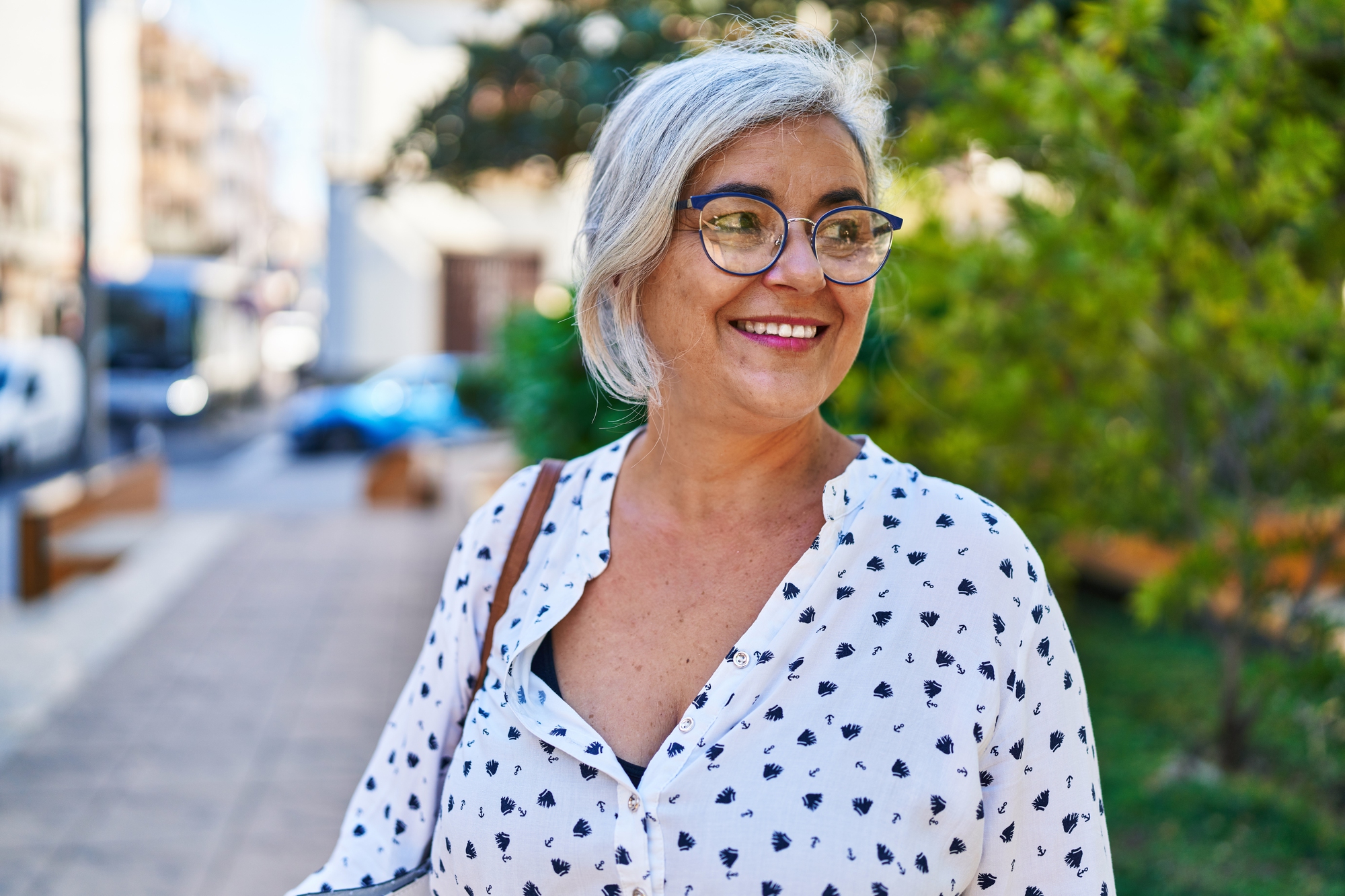 Smiling older woman with gray hair and glasses stands outdoors, wearing a white blouse with black patterns. The background features a tree and a blurred street scene.