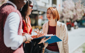 Three people are standing outdoors having a conversation. One person is holding a blue folder. They appear engaged in a lively discussion amid a sunny street with trees and buildings in the background.