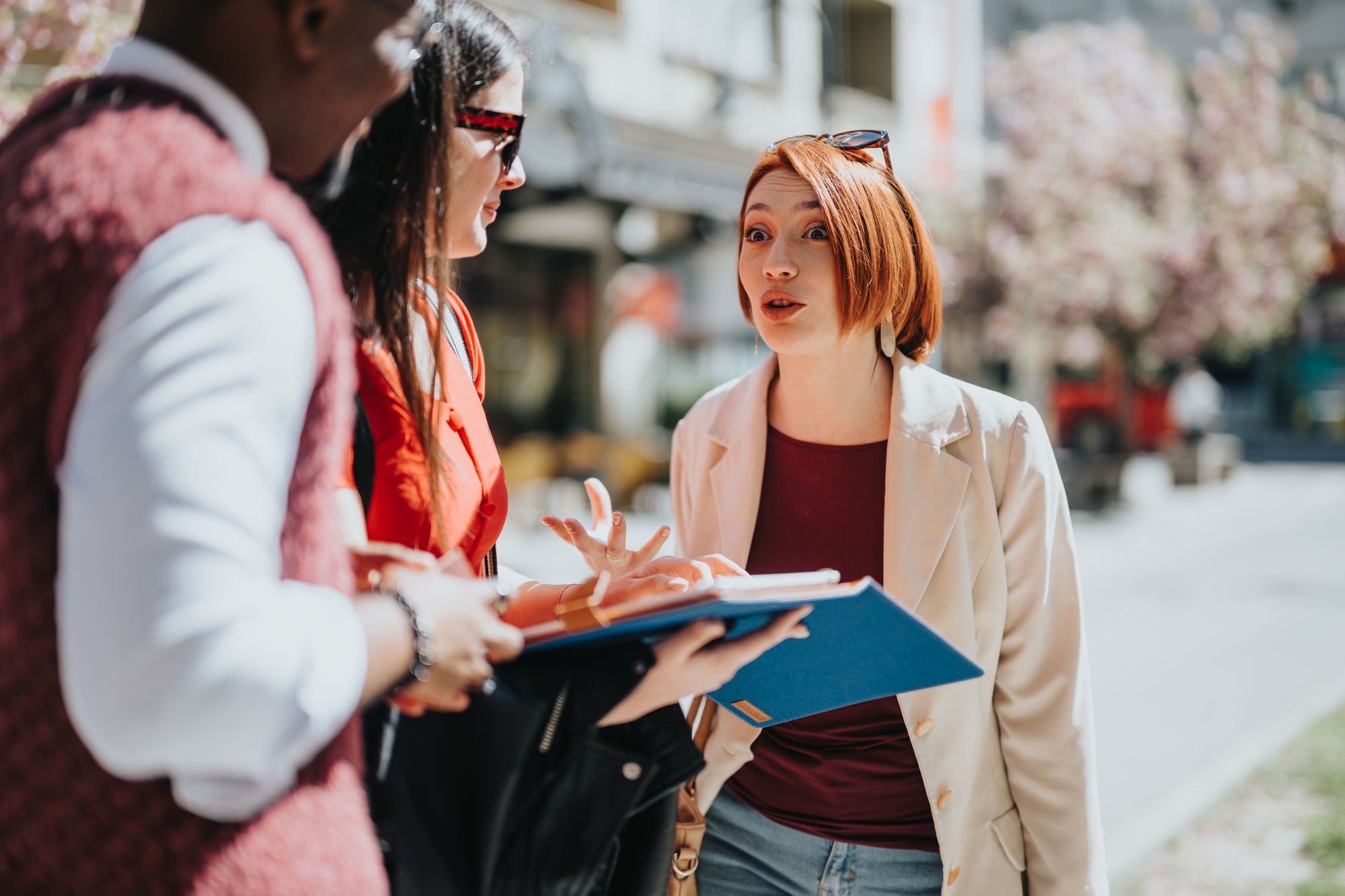 Three people are standing outdoors having a conversation. One person is holding a blue folder. They appear engaged in a lively discussion amid a sunny street with trees and buildings in the background.