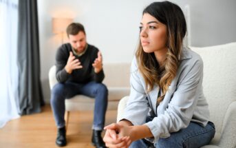 A woman sits on a couch looking upset, with her hands clasped in front. In the background, a man is seated and gesturing as if in conversation. The scene is set in a living room with soft lighting.