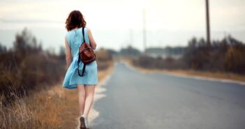 A woman in a light blue dress walks down a rural road, carrying a brown backpack. The scene is serene, with overcast skies and grassy fields on either side of the road.