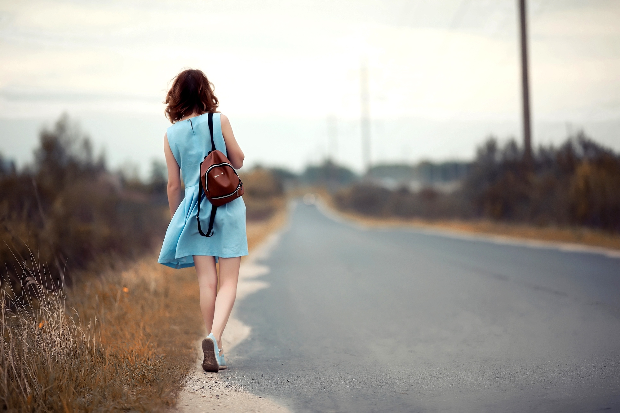 A woman in a light blue dress walks down a rural road, carrying a brown backpack. The scene is serene, with overcast skies and grassy fields on either side of the road.