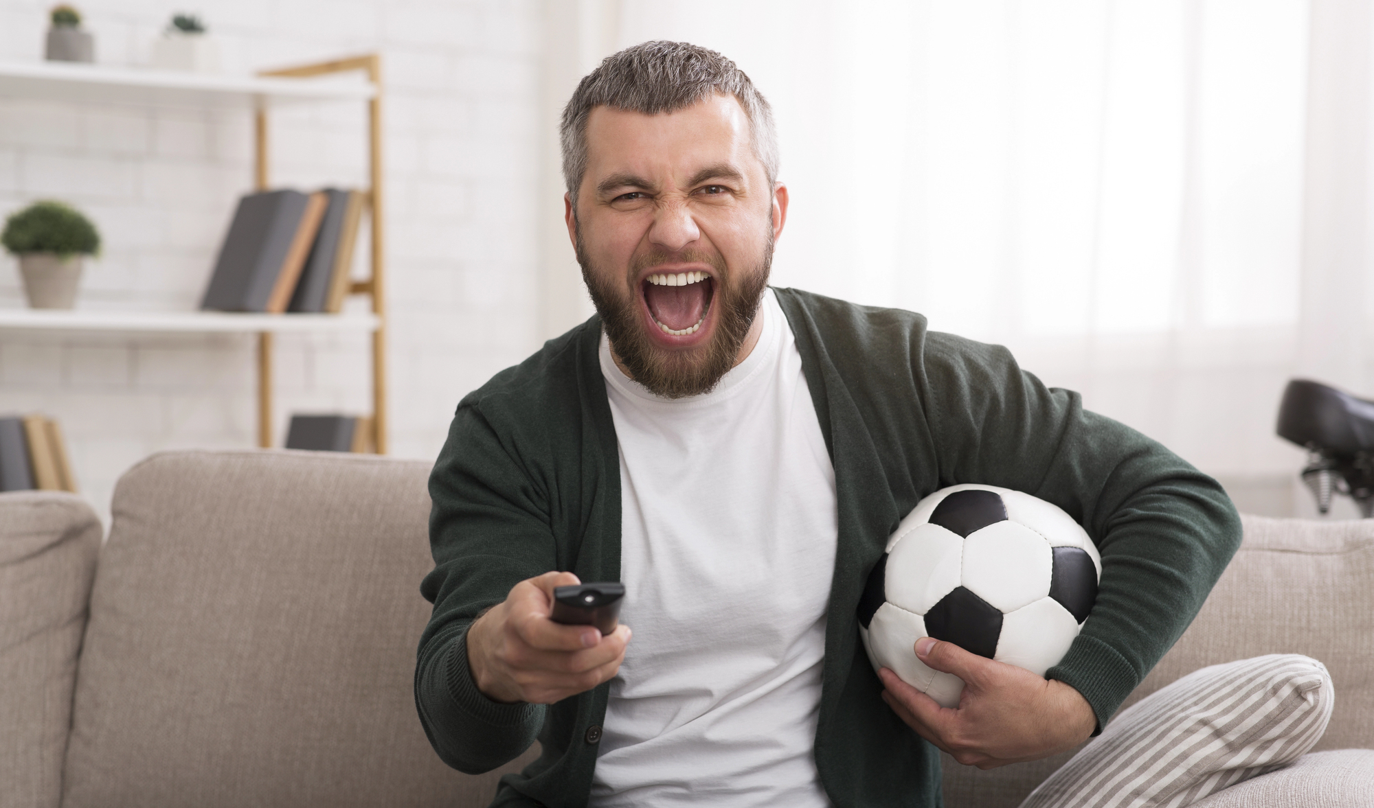 A man with a beard sits on a couch, excitedly cheering while holding a soccer ball under one arm and a remote control in the other hand. Books and a bicycle are in the background.