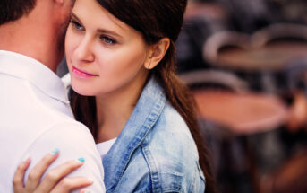 A woman with long dark hair and a denim jacket embraces a man in a white shirt. She gazes thoughtfully into the distance while standing close to him against a blurred background.