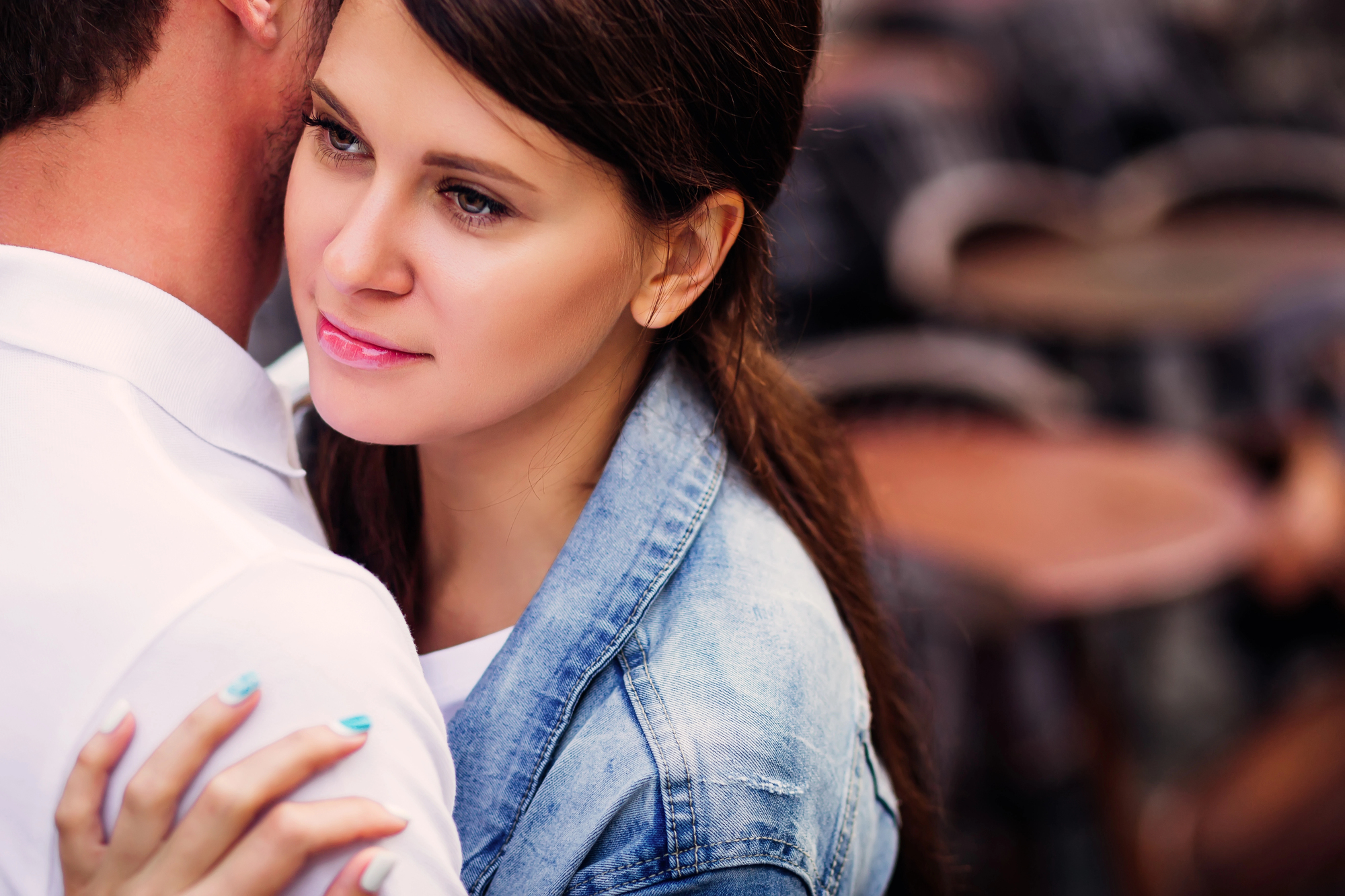 A woman with long dark hair and a denim jacket embraces a man in a white shirt. She gazes thoughtfully into the distance while standing close to him against a blurred background.