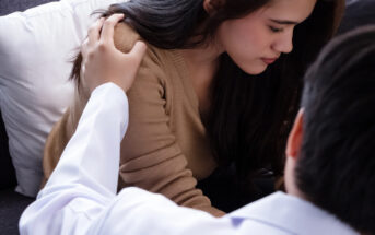 A person in a white shirt sits beside a woman with long dark hair, wearing a brown sweater, on a couch. The person places a comforting hand on her shoulder. Both appear engaged in a serious conversation.