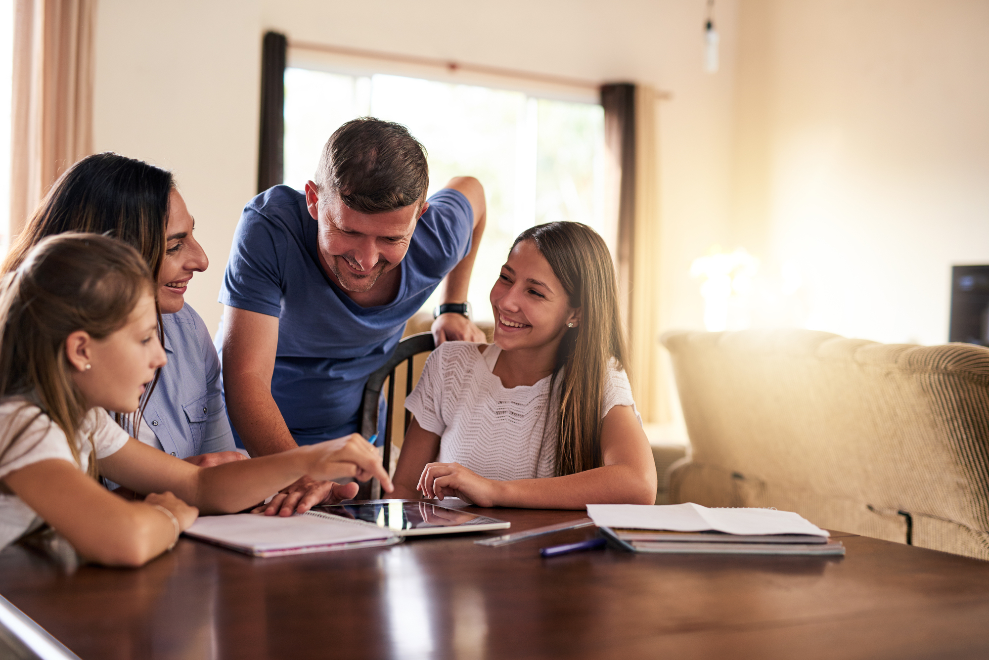 A family of four sits around a table, smiling and looking at a tablet. Two girls, one pointing at the screen, are engaged with their parents who are leaning in. Sunlight filters into the cozy room, creating a warm atmosphere.