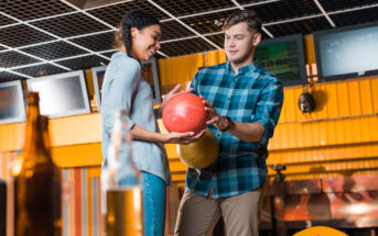 A man and a woman at a bowling alley. The man is holding a red bowling ball, assisting the woman as she looks at it. They are smiling and standing next to each other, with beer bottles visible in the foreground.