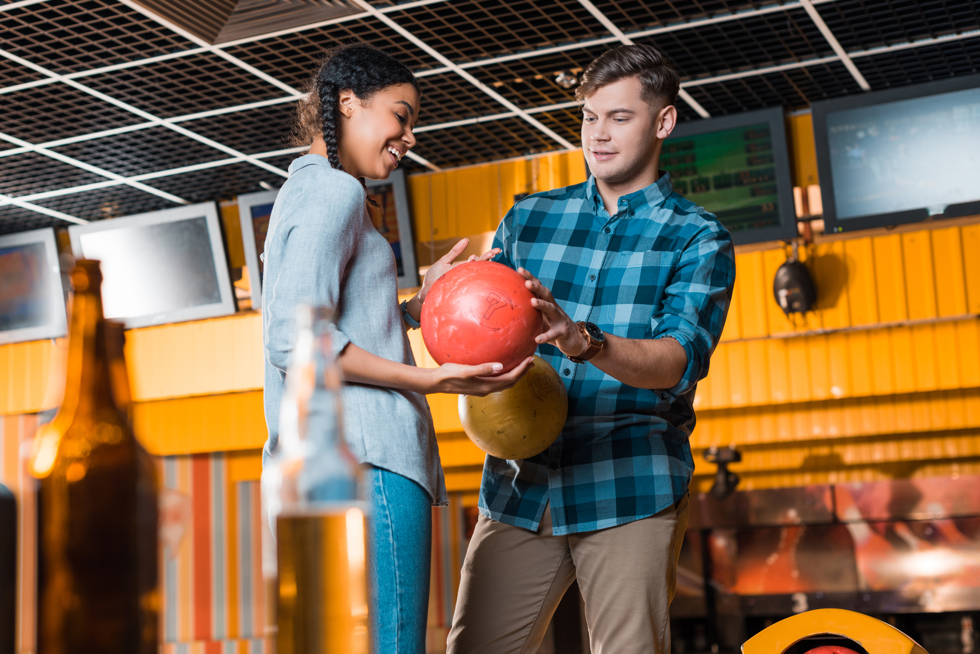 A man and a woman at a bowling alley. The man is holding a red bowling ball, assisting the woman as she looks at it. They are smiling and standing next to each other, with beer bottles visible in the foreground.